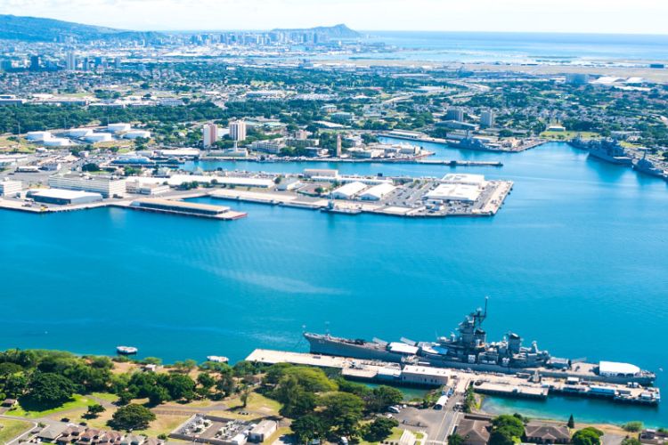 Aerial view of USCG Base Honolulu with Pearl Harbor, docked naval ships, and the Honolulu skyline in the background.