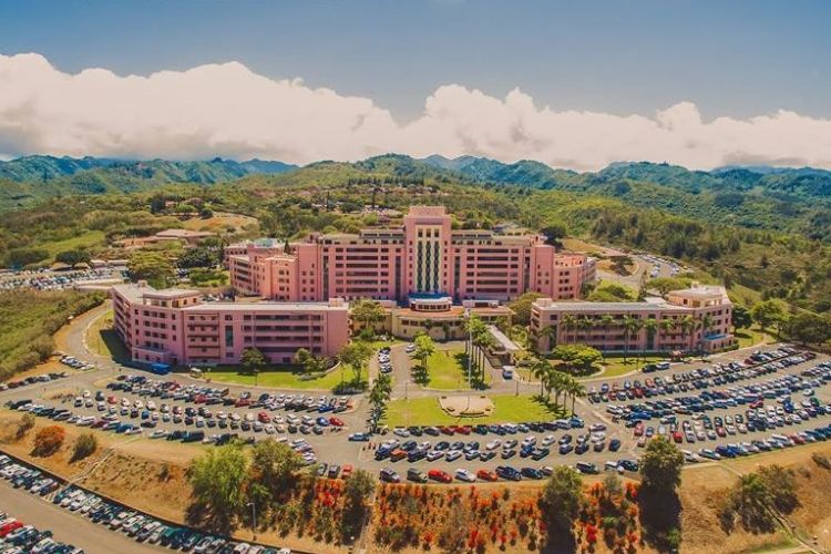 Aerial view of Tripler Army Medical Center, a large pink hospital complex in Hawaii, surrounded by parking lots and lush green mountains.