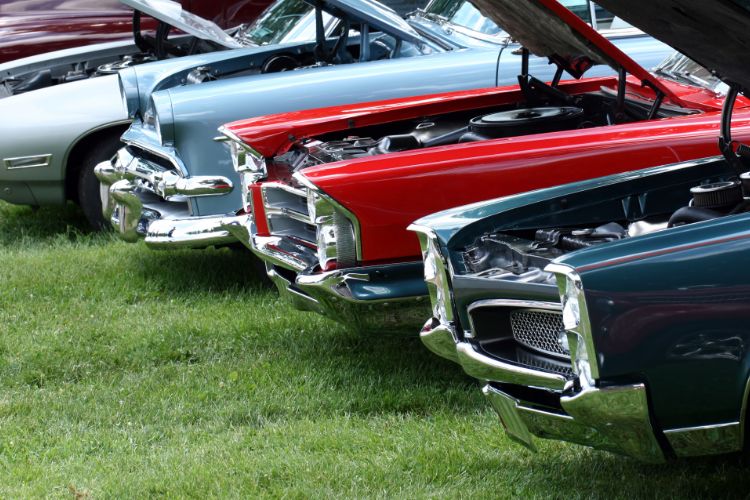 Row of classic cars with open hoods at an outdoor car show, showcasing vintage muscle cars with chrome grilles and powerful engines.